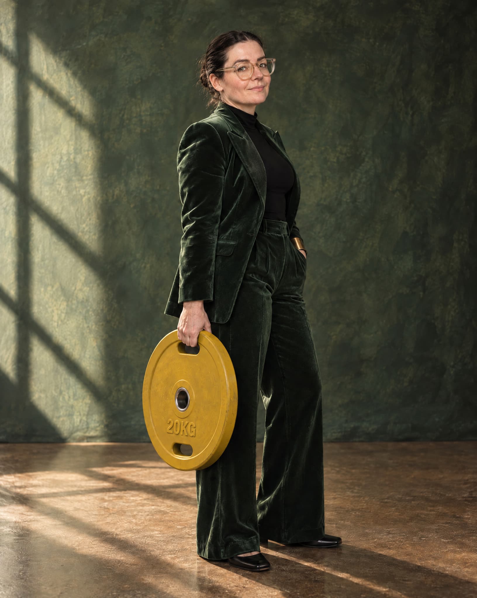 Sandra in a dark green velvet suit, holding a 20 kg weight plate at her side.